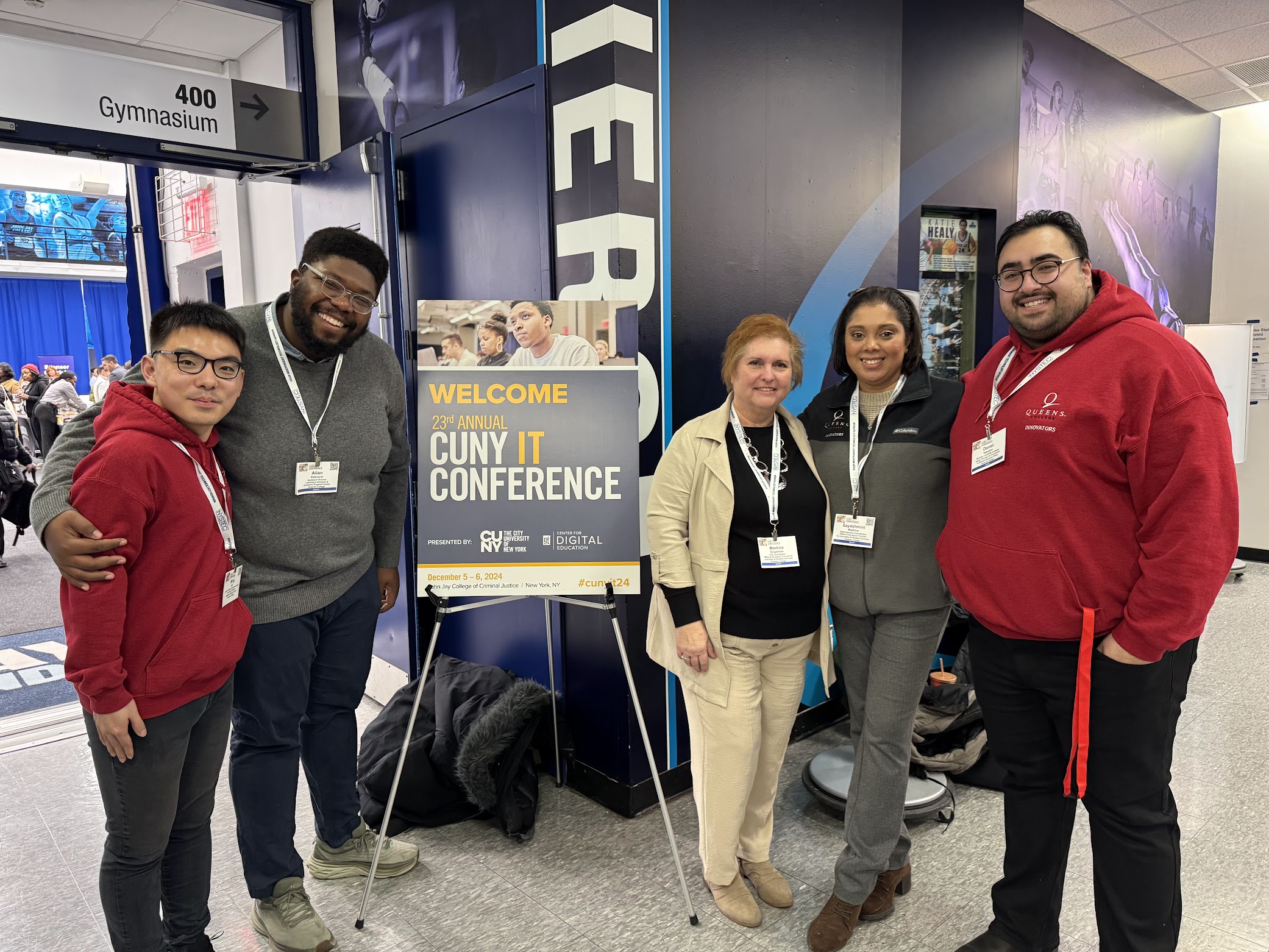 A group of 5 people standing next to a CUNY IT Conference poster